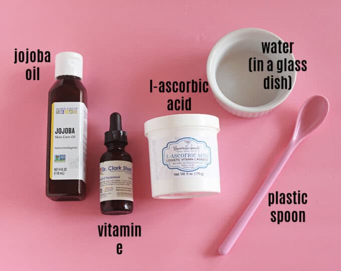Vitamin C powder, jojoba oil, pink spoon, and white ramekin on a pink background.