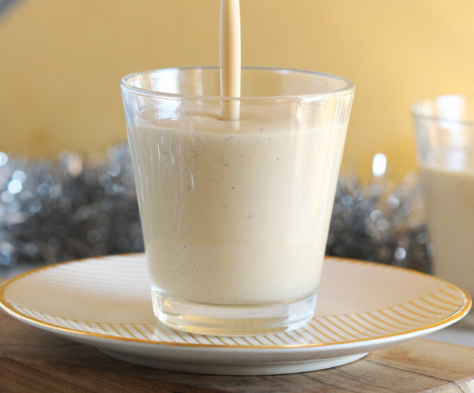Pouring eggnog into a clear glass on a plate.