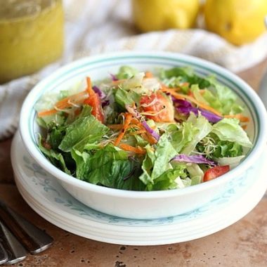 A bowl of salad and vegetables sitting on a table with two lemons, plates, silverware, and a jar of salad dressing.