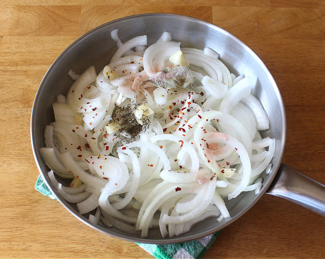 Sliced onions in a steel saute pan with thyme and red pepper flakes on top.