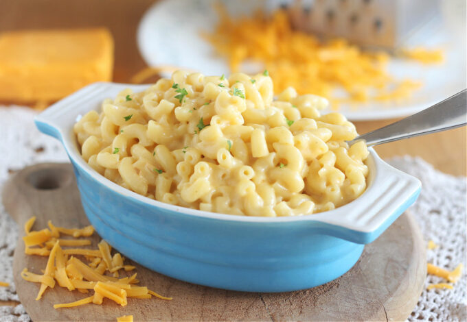 Macaroni and cheese in a small blue baking crock with cheddar cheese around.