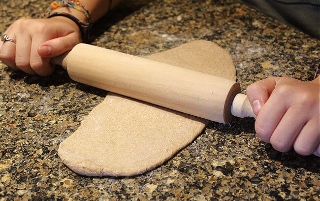 Hands rolling out dough into an oval shape on a granite countertop.