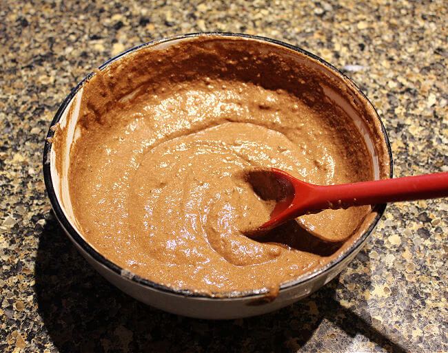 Chocolate batter in a large white bowl on a granite counter with a red spoon.