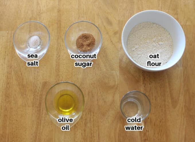 Oat flour, sugar, salt, oil, and water laid out in bowls on a wood table.