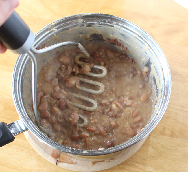 Pinto beans being mashed with a potato masher in a pot.