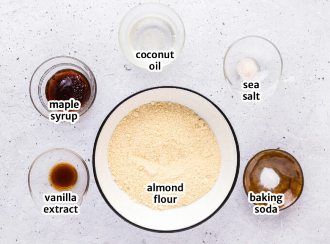Ingredients for cookies, including almond flour, maple syrup, and baking soda, laid out on a table.
