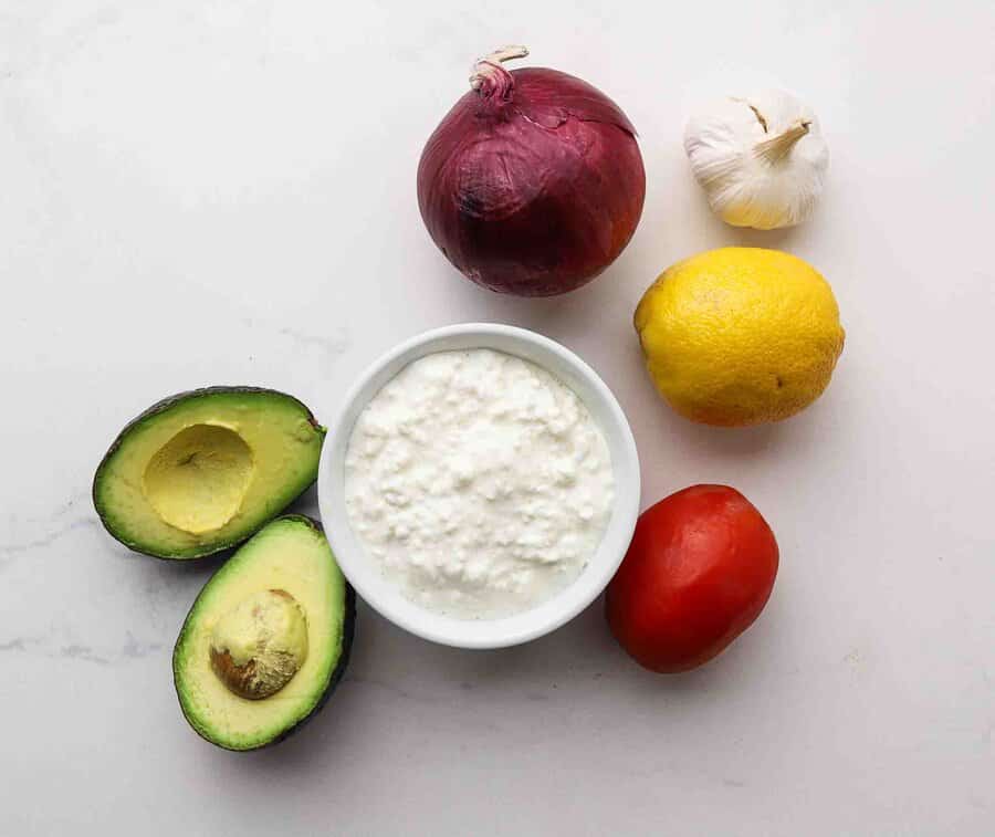 Cottage cheese, avocado, onion, lemon, garlic, and tomato on a table.