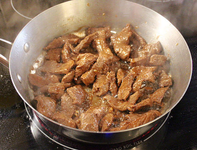 Caramelized meat cooking in a large stainless steel pan.