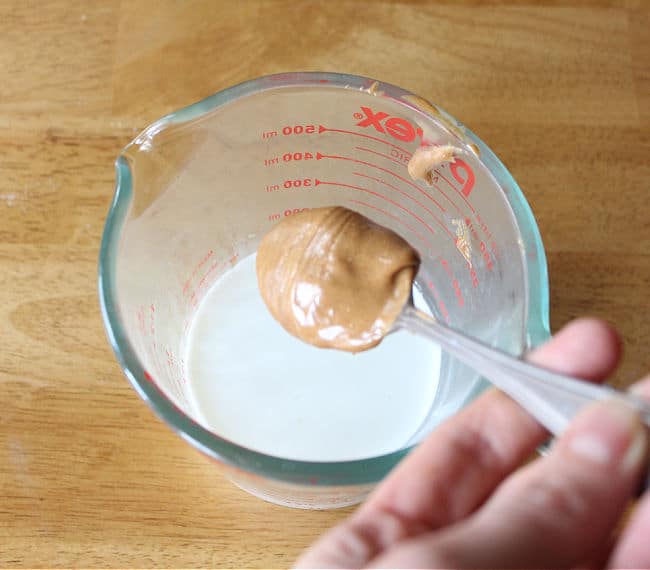 Spoonful of peanut butter being mixed into a measuring cup full of milk.