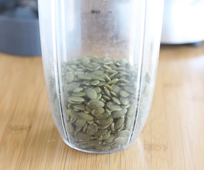 Green pumpkin seeds in a plastic blender cup on a wood counter.