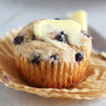 Blueberry muffin topped with butter on a stack of paper muffin cups.